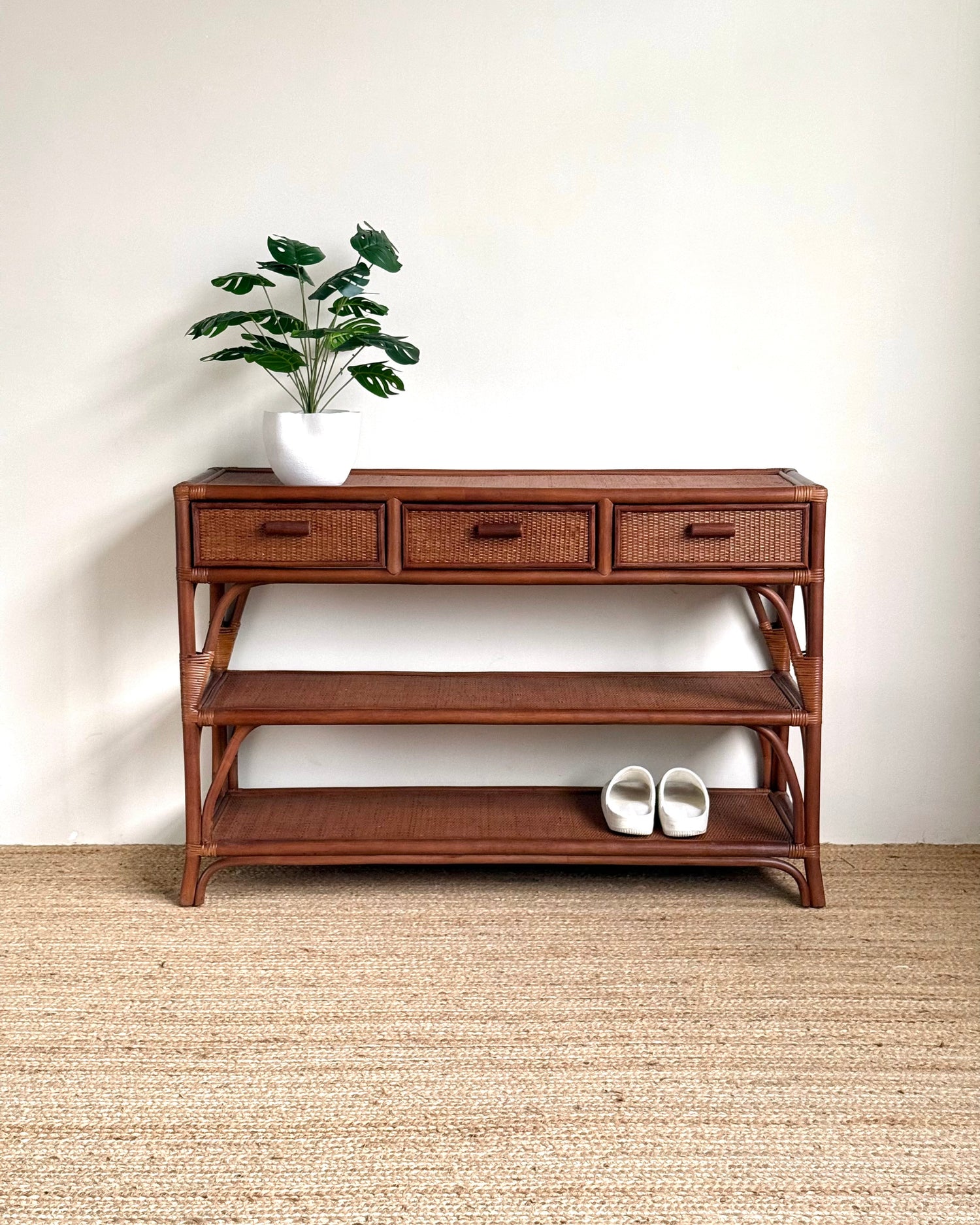 A mid-century modern wooden console table with three drawers, open shelves, and a woven rattan frame, styled with a potted plant and white slip-on shoes against a white wall and jute rug.