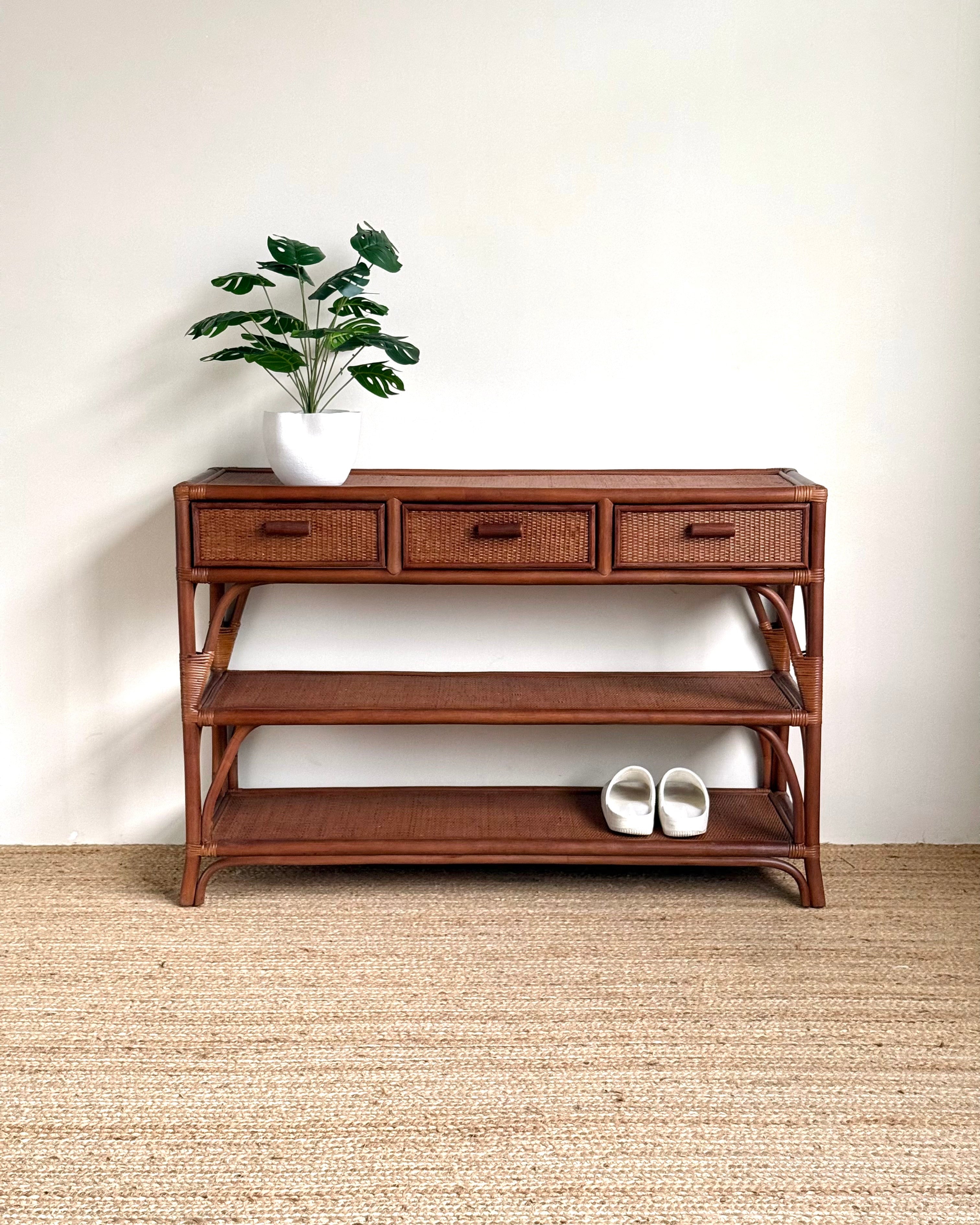 A mid-century modern wooden console table with three drawers, open shelves, and a woven rattan frame, styled with a potted plant and white slip-on shoes against a white wall and jute rug.