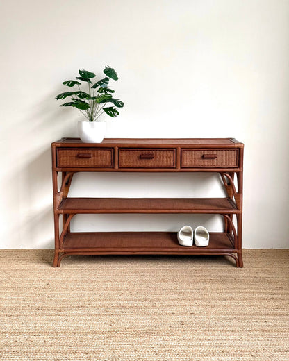 A mid-century modern wooden console table with three drawers, open shelves, and a woven rattan frame, styled with a potted plant and white slip-on shoes against a white wall and jute rug.