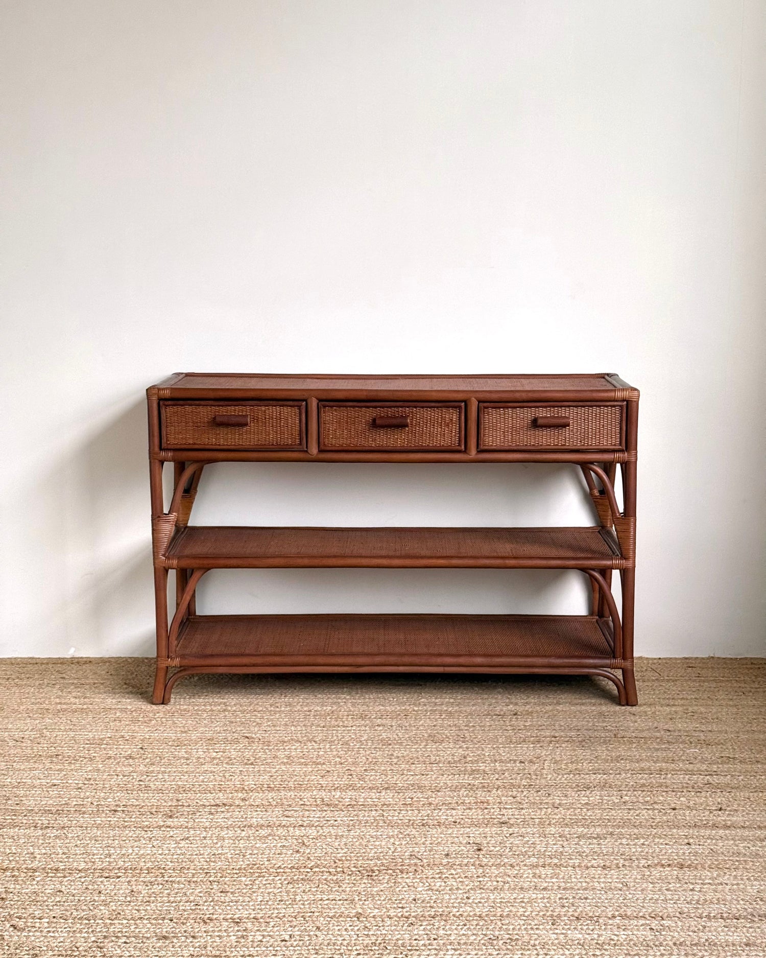A mid-century modern wooden console table with three drawers, woven rattan drawer fronts, an open middle shelf, and a lower shelf, set against a plain white wall and light beige woven rug.