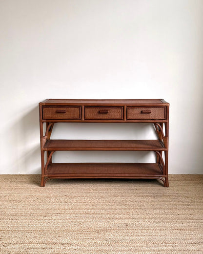 A mid-century modern wooden console table with three drawers, woven rattan drawer fronts, an open middle shelf, and a lower shelf, set against a plain white wall and light beige woven rug.