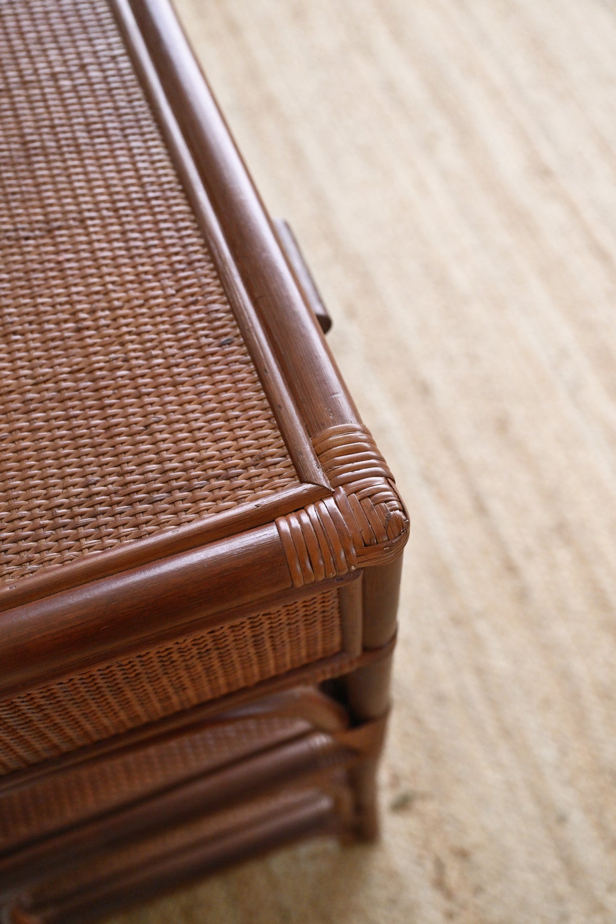 Close-up of a brown rattan or wicker furniture piece with a woven top and carved wooden corner details, set on a light beige textured floor.