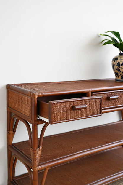 A brown rattan console table with woven drawer fronts and wooden handles, shown against a plain white wall with a potted plant in the background.