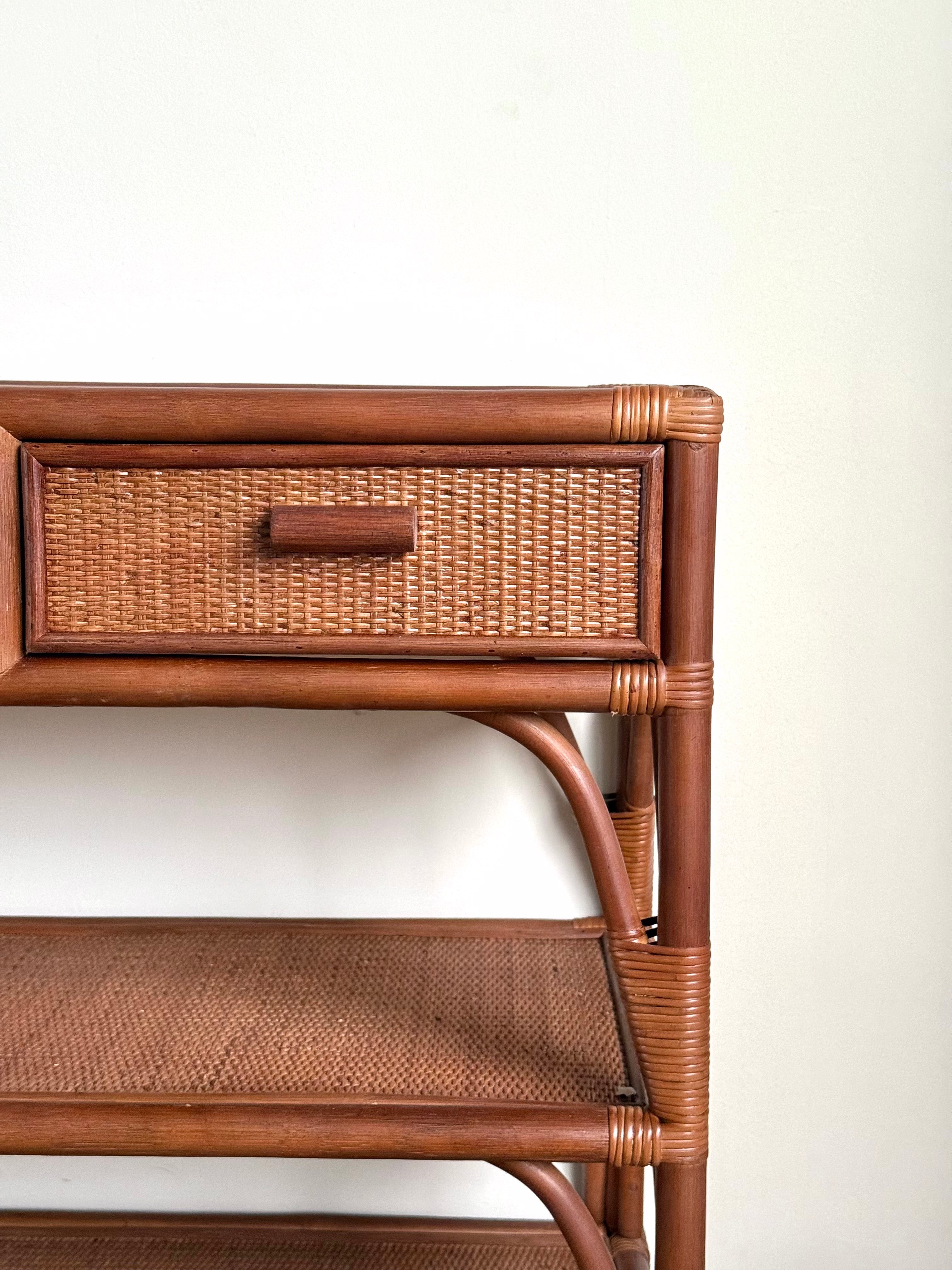 Close-up of a brown rattan and wicker storage shelf with a single drawer and woven shelf, set against a plain off-white wall.
