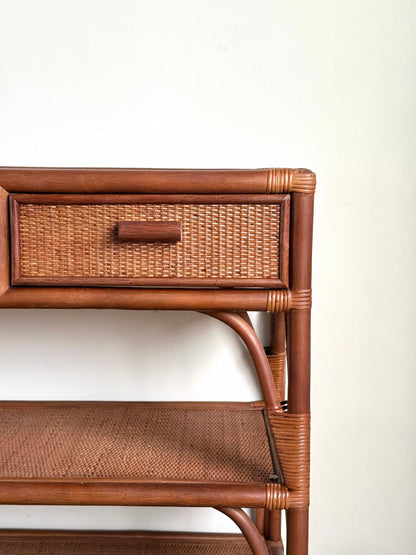 Close-up of a brown rattan and wicker storage shelf with a single drawer and woven shelf, set against a plain off-white wall.