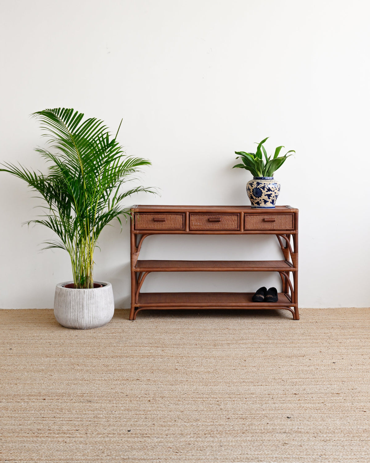 A mid-century modern wooden console table with three drawers and open shelves, styled in a minimalist room with a beige jute rug, a potted palm, and a small potted plant on top.