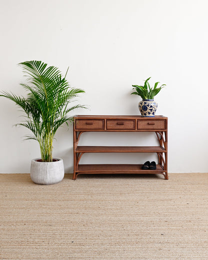 A mid-century modern wooden console table with three drawers and open shelves, styled in a minimalist room with a beige jute rug, a potted palm, and a small potted plant on top.