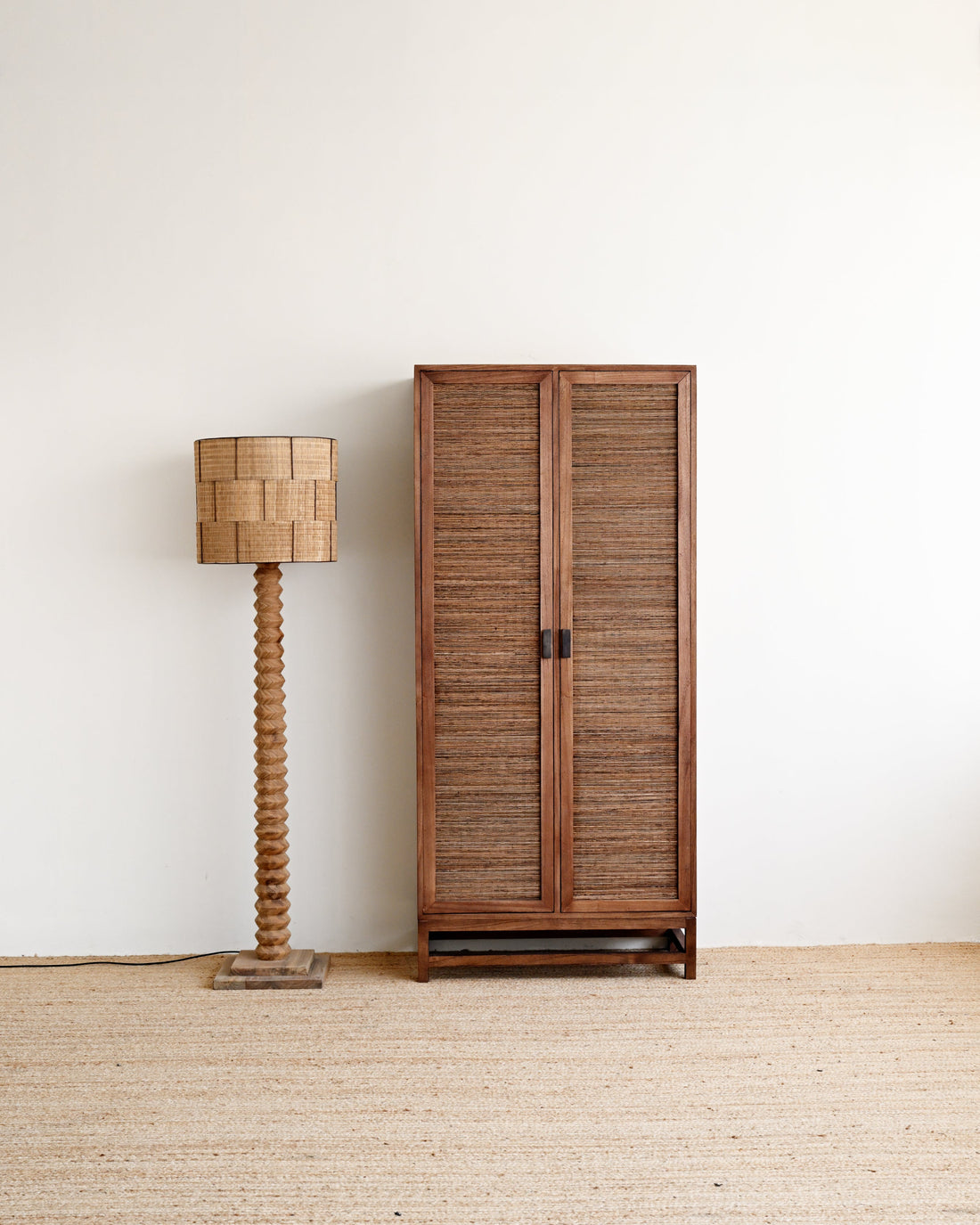 A tall wooden two-door cabinet with woven slat doors stands against a plain white wall on a light beige woven floor, next to a matching woven floor lamp.
