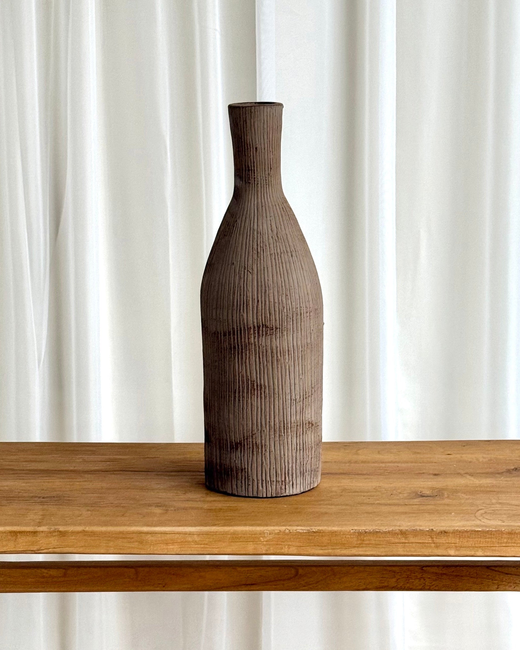 A tall, narrow ceramic vase with a textured, vertically ribbed surface in a dark brown finish, displayed on a light wood table against a backdrop of white curtains.