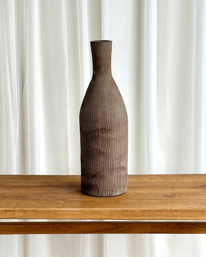 A tall, narrow ceramic vase with a textured, vertically ribbed surface in a dark brown finish, displayed on a light wood table against a backdrop of white curtains.