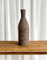 A tall, narrow ceramic vase with a textured, vertically ribbed surface in a dark brown finish, displayed on a light wood table against a backdrop of white curtains.