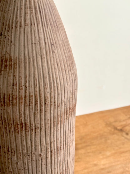 A close-up of a ribbed ceramic vase with a textured, distressed finish in warm beige and brown tones, standing on a wooden surface against a plain off-white wall.