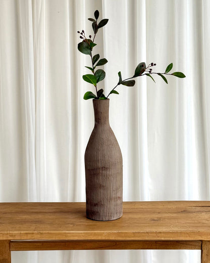 A tall ribbed ceramic vase in a warm taupe-brown color, holding a green leafy sprig, set on a rustic wooden table against a white curtain background.