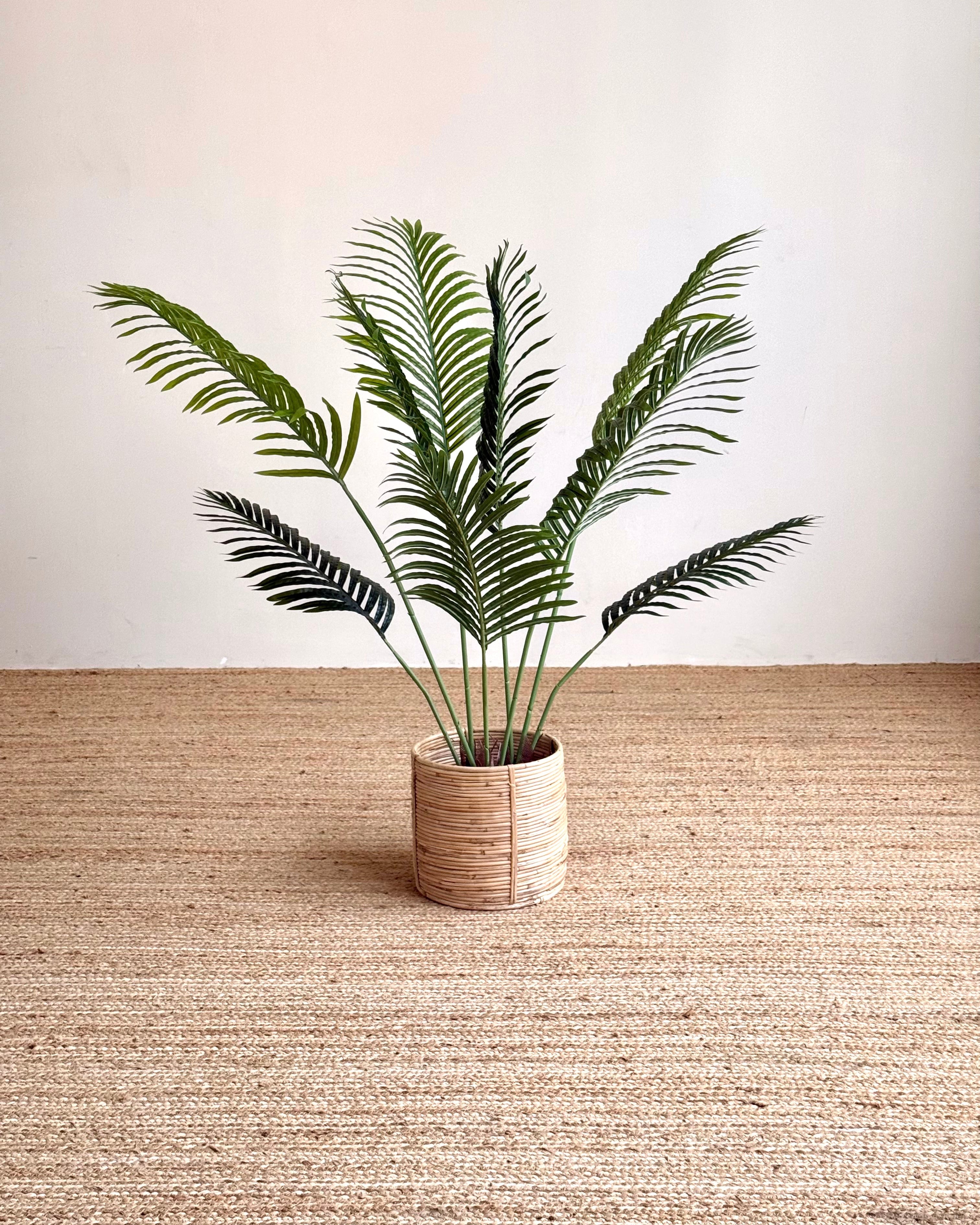 A potted artificial palm plant with green fronds in a cylindrical woven beige pot, placed on a textured beige rug against a plain off-white wall.