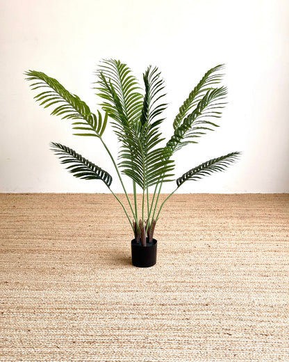 An artificial green palm frond plant in a small black pot, set against a neutral beige woven rug and plain white wall background.