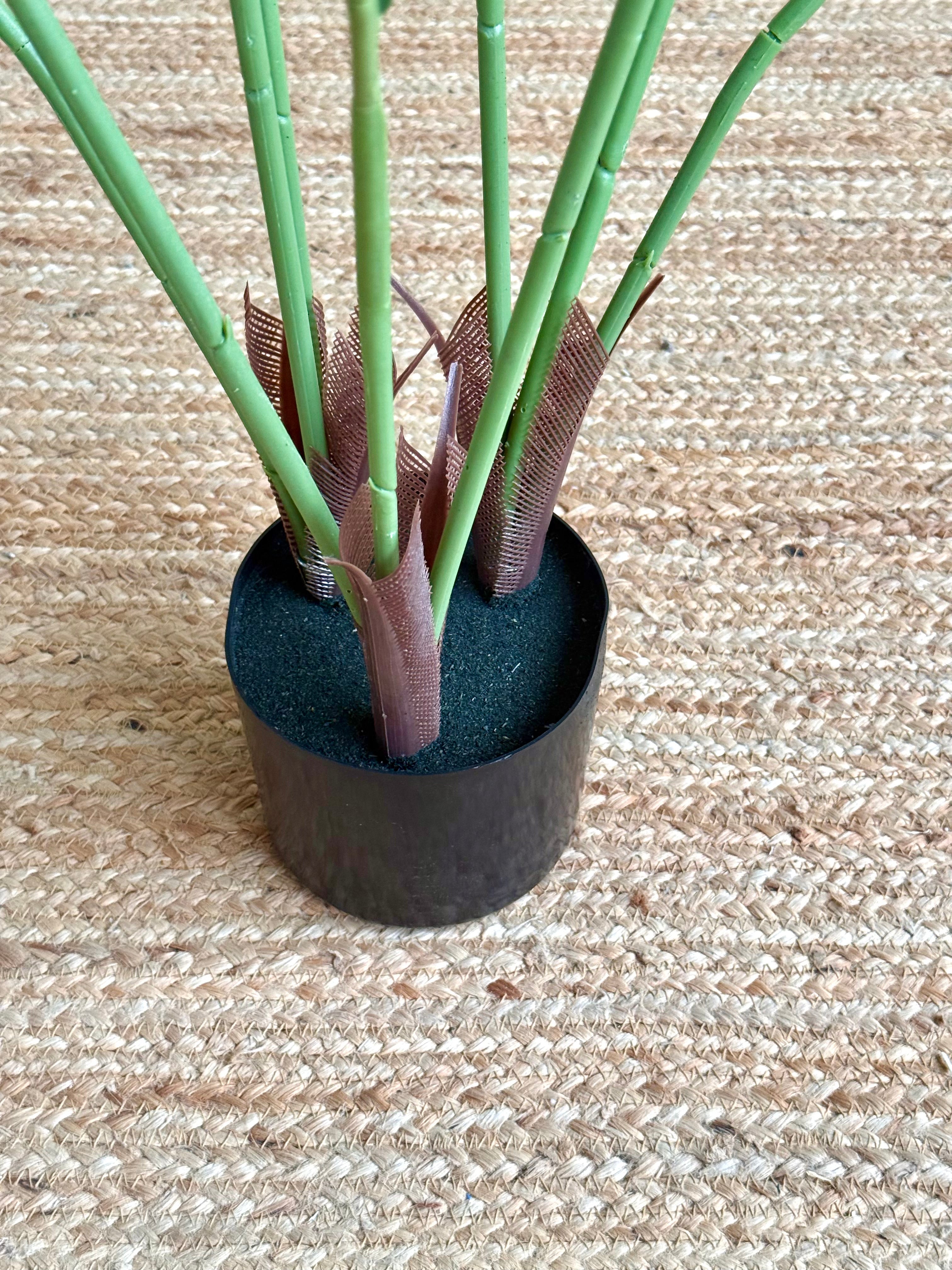 A potted artificial plant with long light green stems and brown faux leaves, set in a black plastic pot on a woven beige rug.