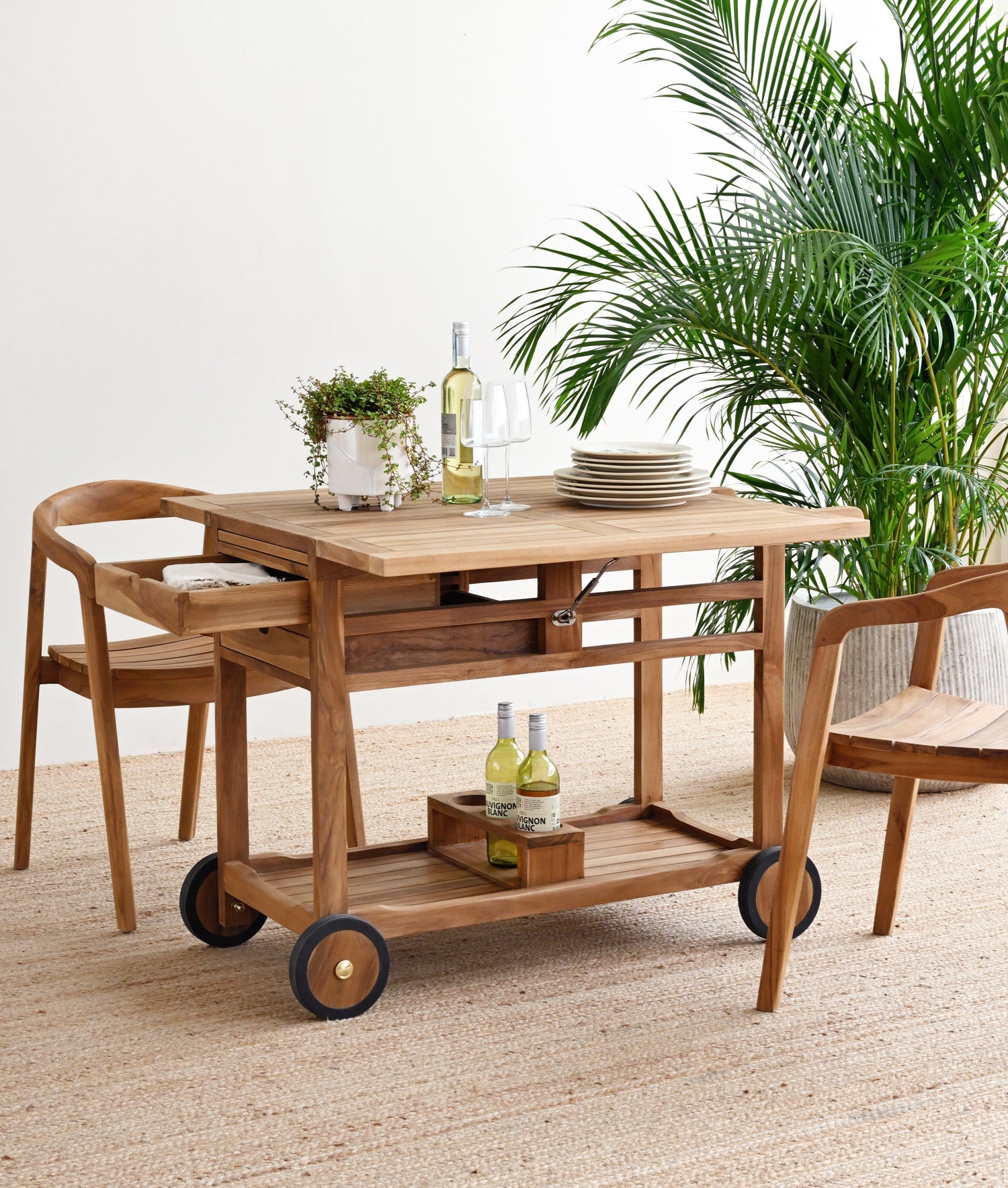 Wooden serving cart with drinks and plates on a beige carpet, surrounded by plants.