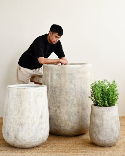 Person sitting on a large concrete planter with smaller planters and a plant on a wooden floor.