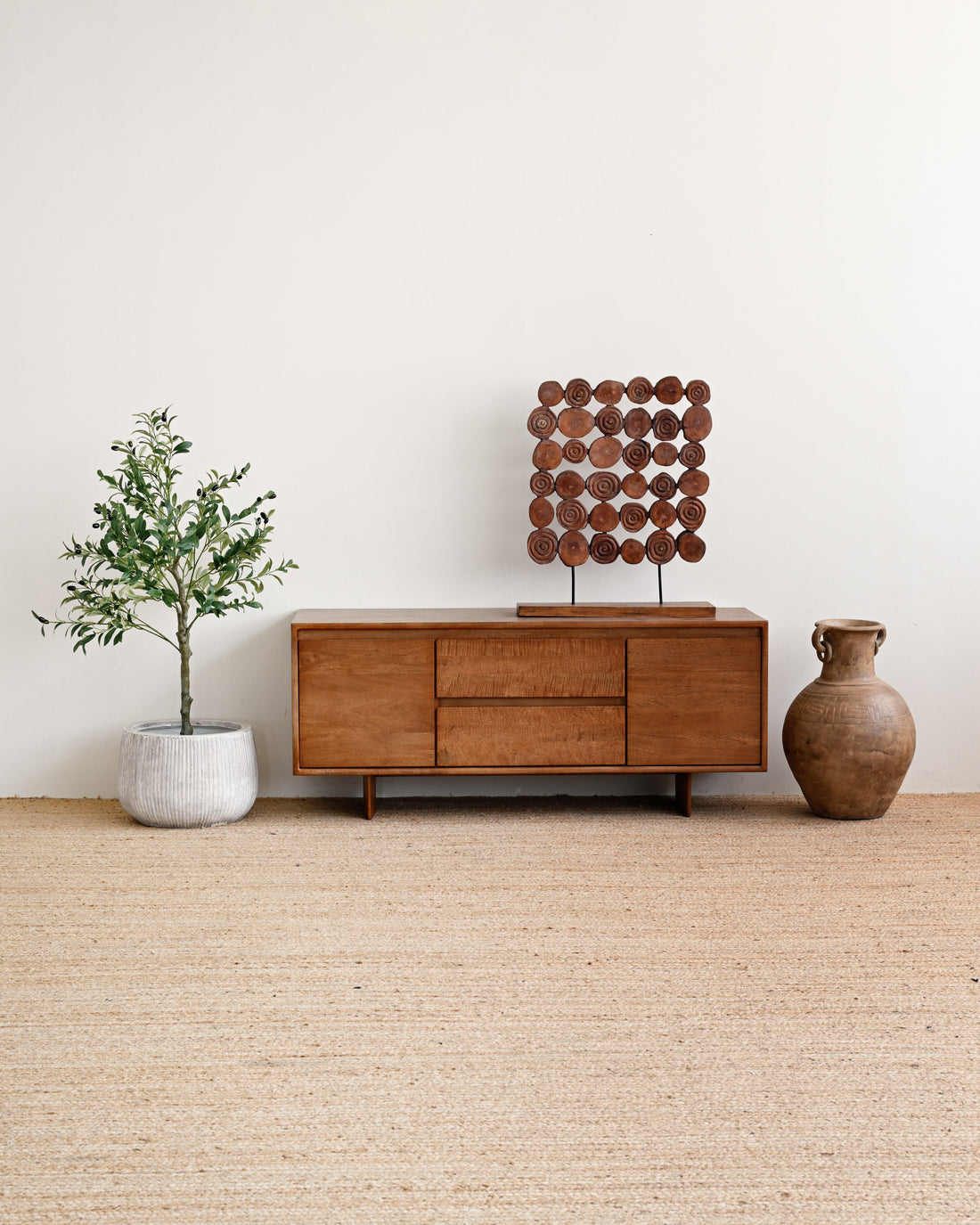 A mid-century modern wooden sideboard cabinet with a geometric wooden sculpture on top, styled in a minimalist room with a beige jute rug, a potted plant, and a ceramic vase against a white wall.