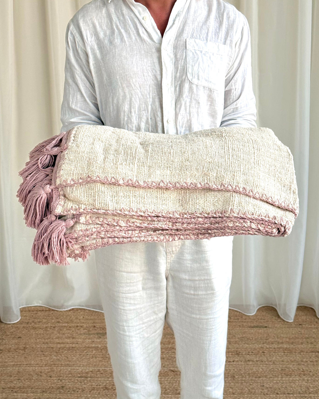 A person wearing a white linen shirt and white pants holds a folded cream-colored woven throw blanket with dusty pink fringe tassels and textured stripes.