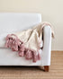 A textured throw blanket in cream and dusty pink with tassel fringe, draped over the arm of a white upholstered armchair on a woven rug.