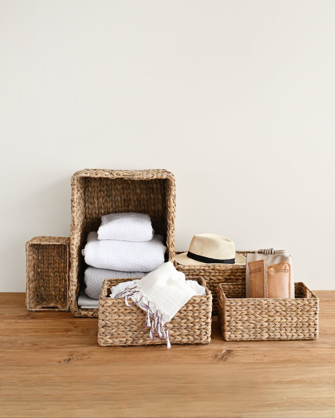 A set of natural woven wicker storage baskets in different sizes, styled on a wooden tabletop against a plain off-white wall. The baskets are shown holding folded white towels, a white throw blanket with fringe, a beige straw hat, and a brown paper bag.