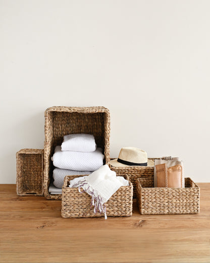 A set of natural woven wicker storage baskets in different sizes, styled on a wooden tabletop against a plain off-white wall. The baskets are shown holding folded white towels, a white throw blanket with fringe, a beige straw hat, and a brown paper bag.