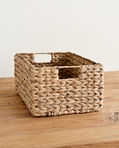 A rectangular woven storage basket made from natural seagrass or wicker, featuring integrated handles, resting on a light wood surface against a plain off-white wall.