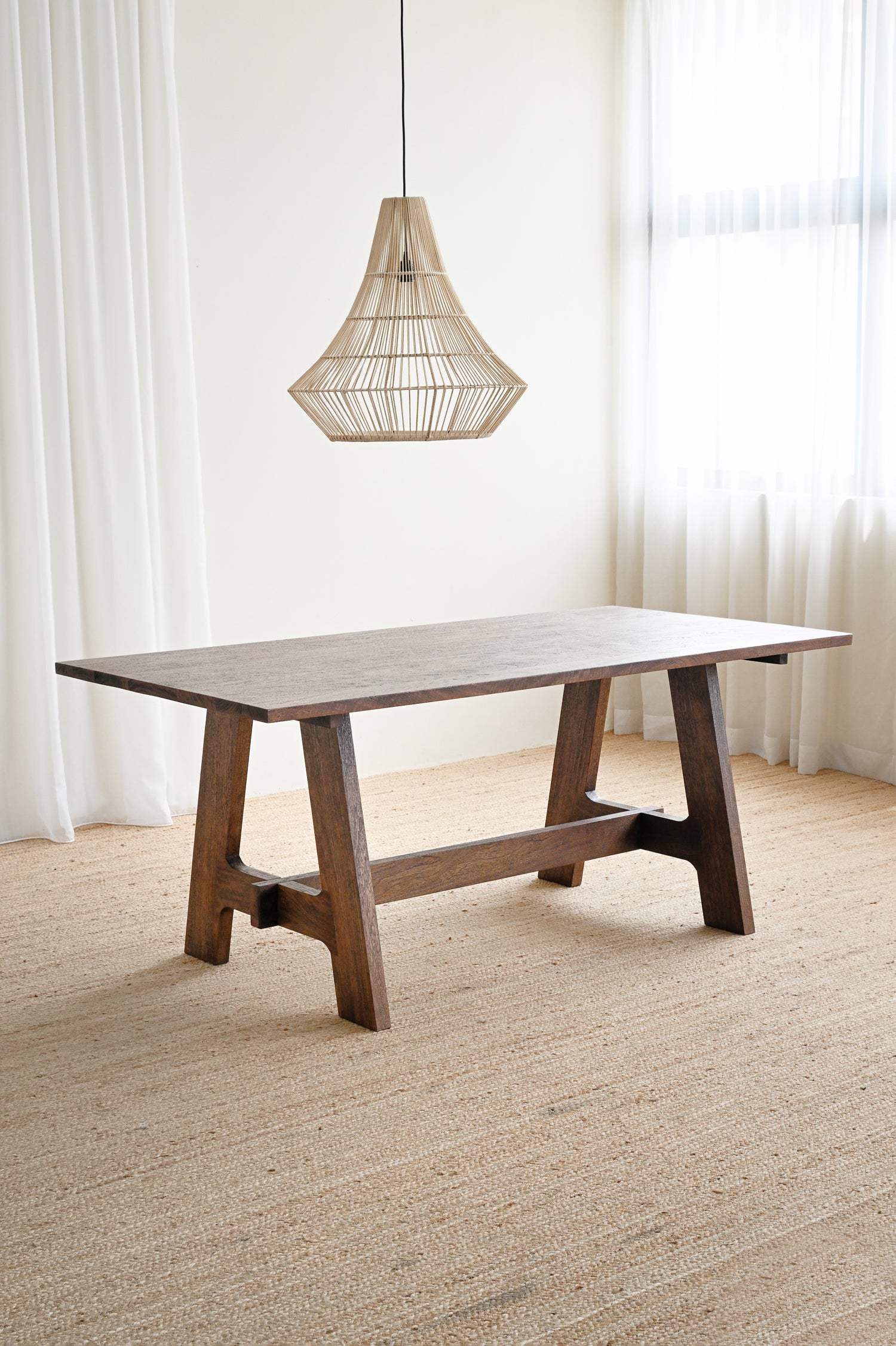 Wooden dining table with a hanging light fixture in a room with white curtains and beige carpet.