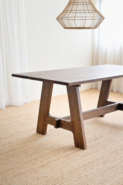 Wooden dining table with a textured surface in a room with white curtains and a woven lampshade.