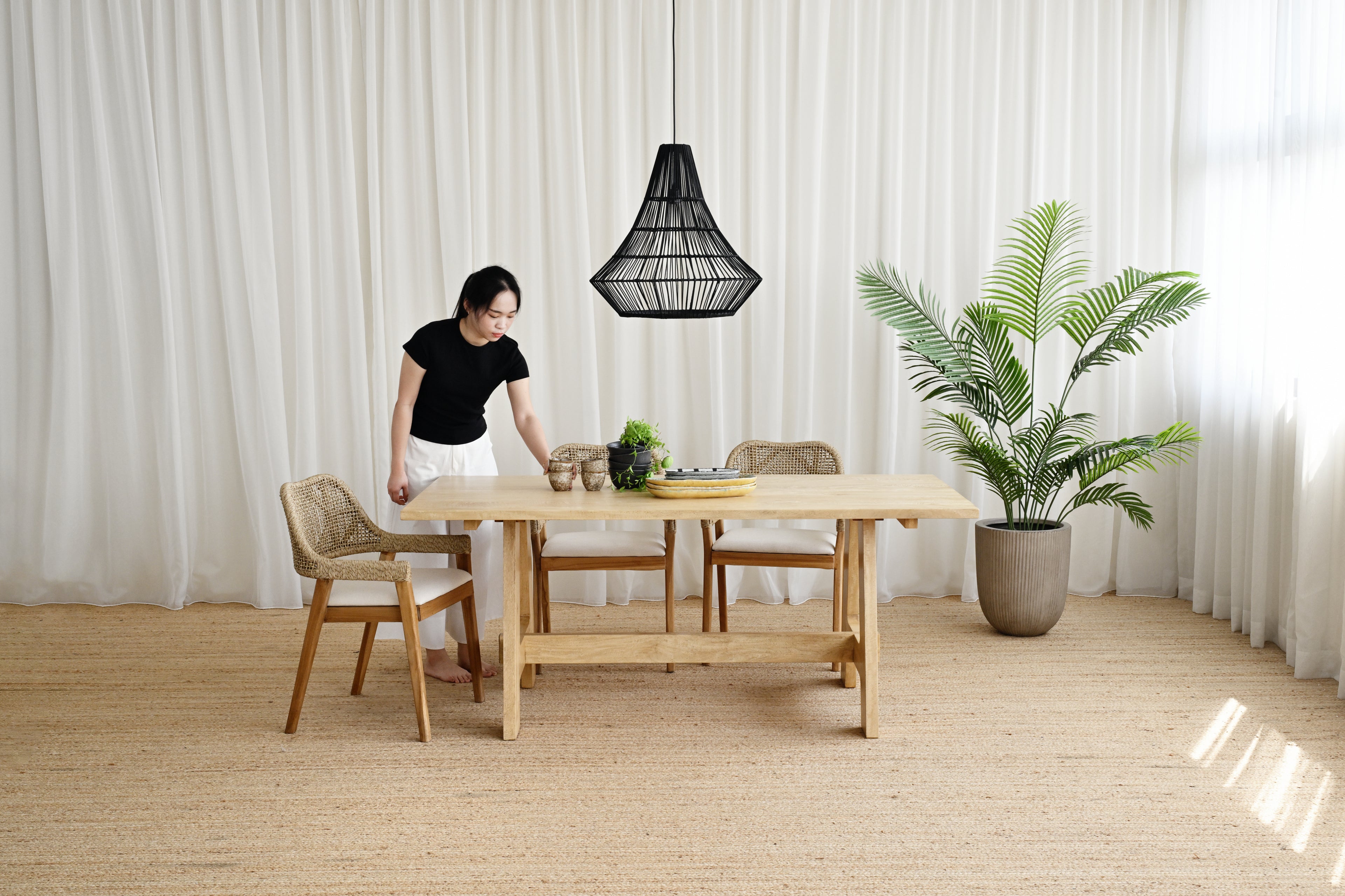 Woman standing by a wooden dining table in a room with white curtains and a potted plant.