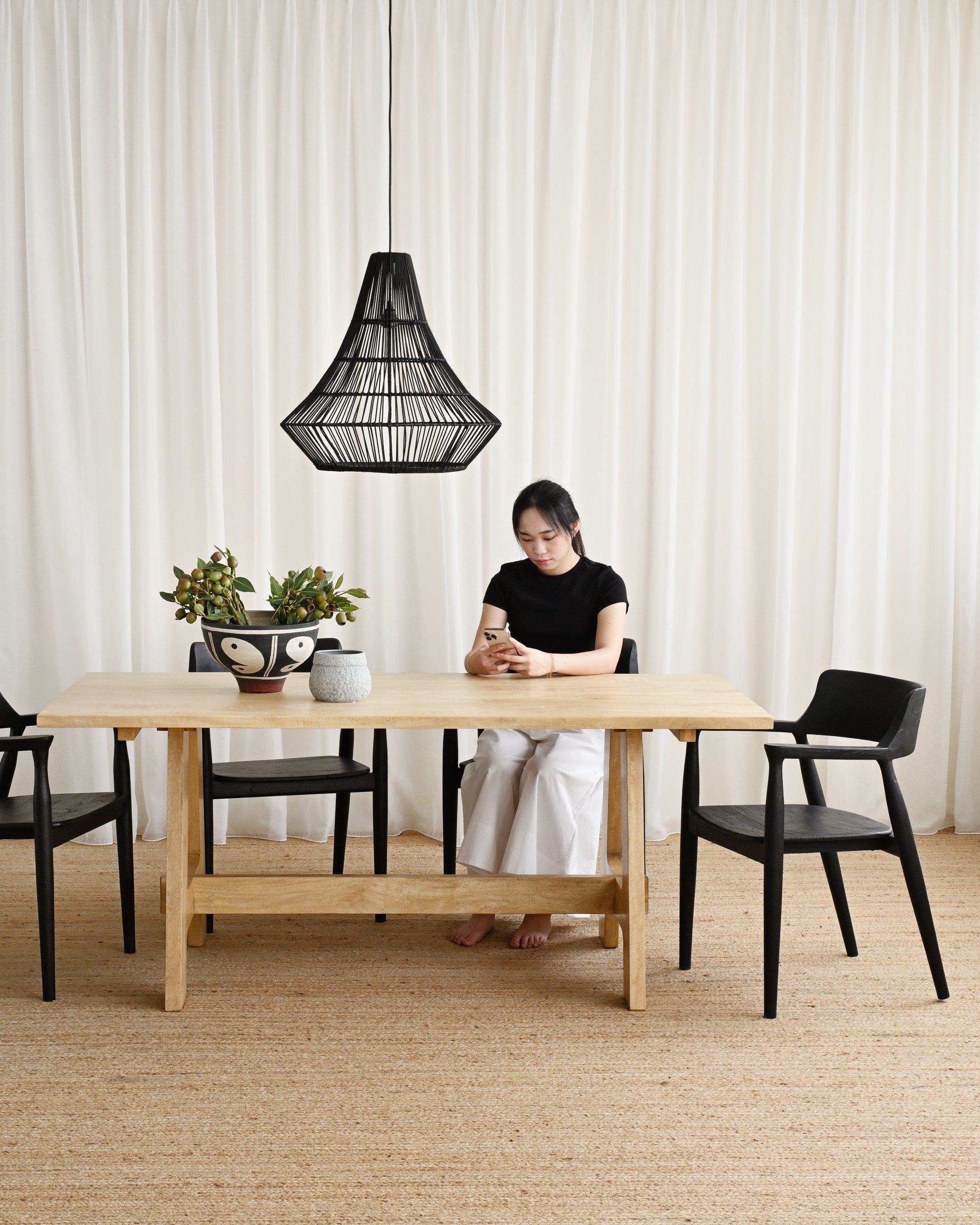 Person sitting at a wooden table with black chairs in a room with white curtains.