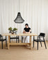 Person sitting at a wooden table with black chairs in a room with white curtains.