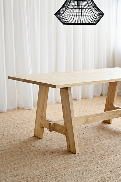 Wooden dining table with a black lampshade in a room with white curtains and a textured floor.