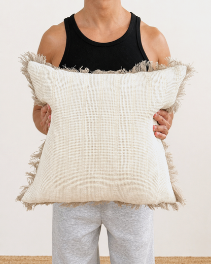 Person holding a large decorative pillow with a textured pattern and frayed edges.