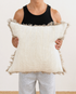 Person holding a large decorative pillow with a textured pattern and frayed edges.