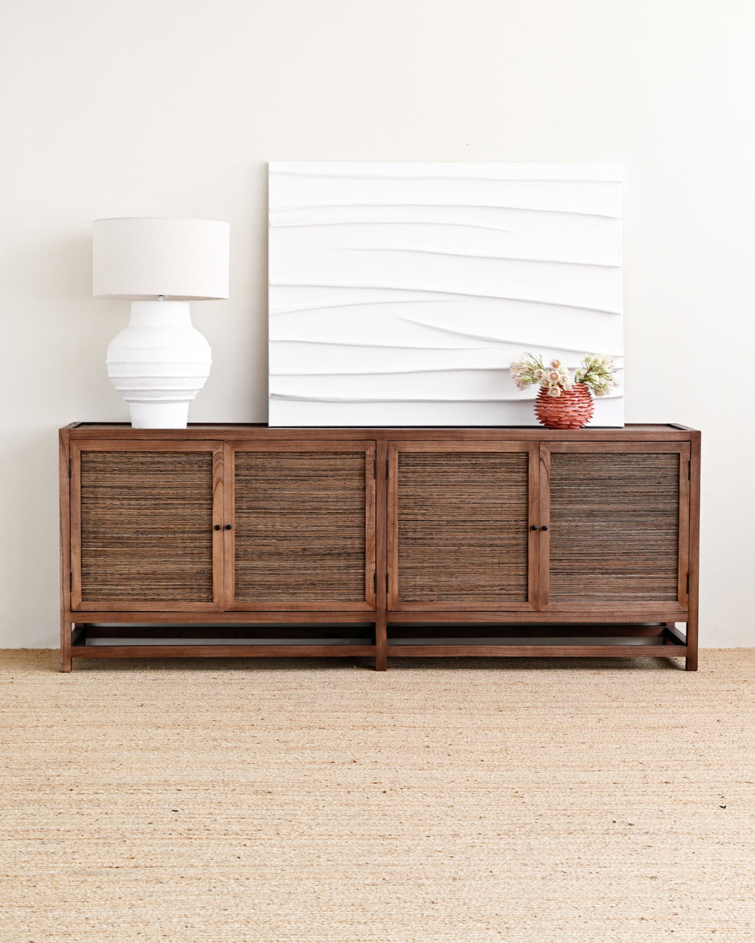 A dark brown wooden sideboard with woven rattan-style doors, styled in a minimalist room with a white table lamp, a white textured canvas print, and a small potted plant on top, set against a light beige wall and jute rug.