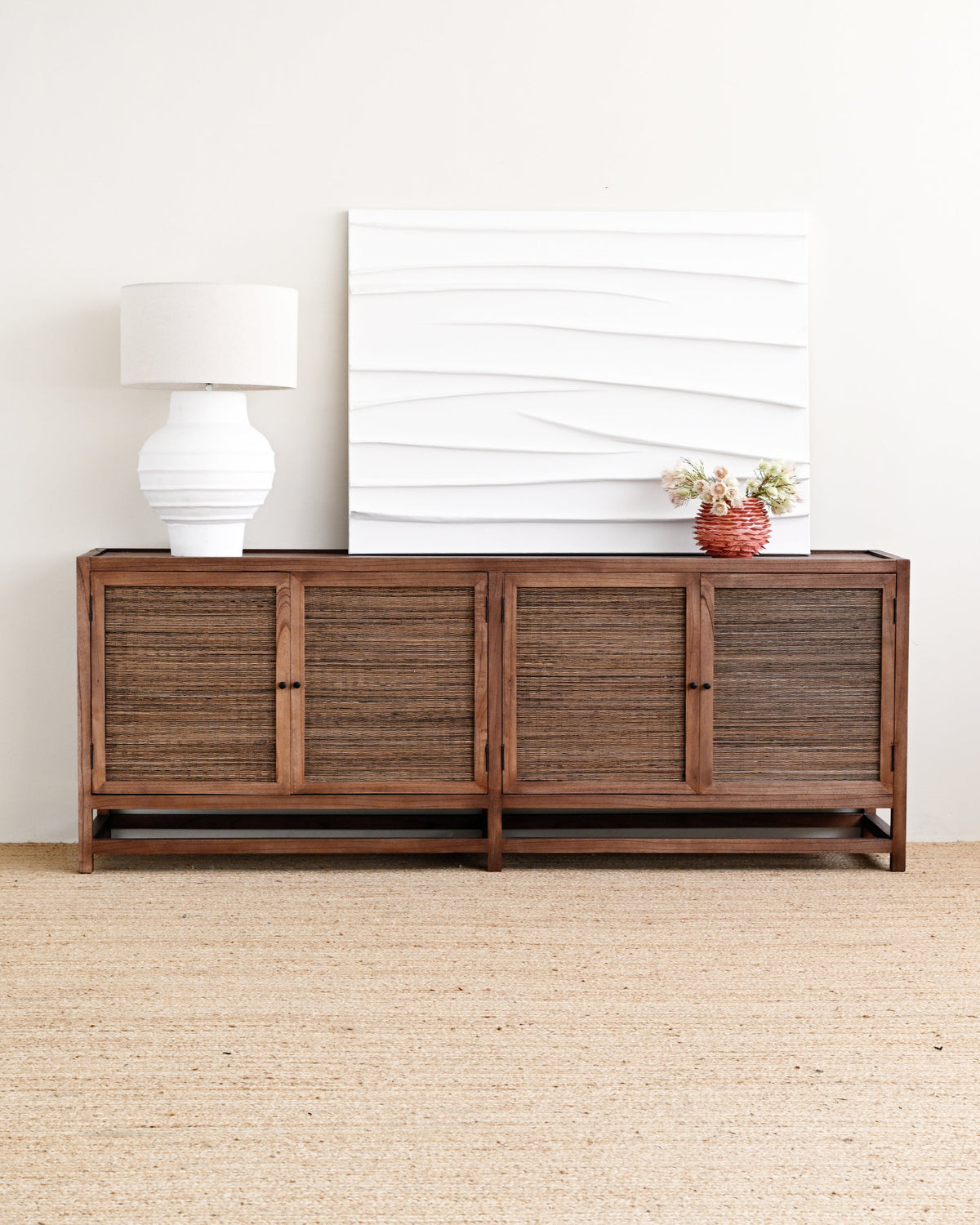 A dark brown wooden sideboard with woven rattan-style doors, styled in a minimalist room with a white table lamp, a white textured canvas print, and a small potted plant on top, set against a light beige wall and jute rug.