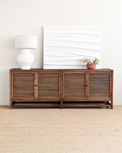 A dark brown wooden sideboard with woven rattan-style doors, styled in a minimalist room with a white table lamp, a white textured canvas print, and a small potted plant on top, set against a light beige wall and jute rug.