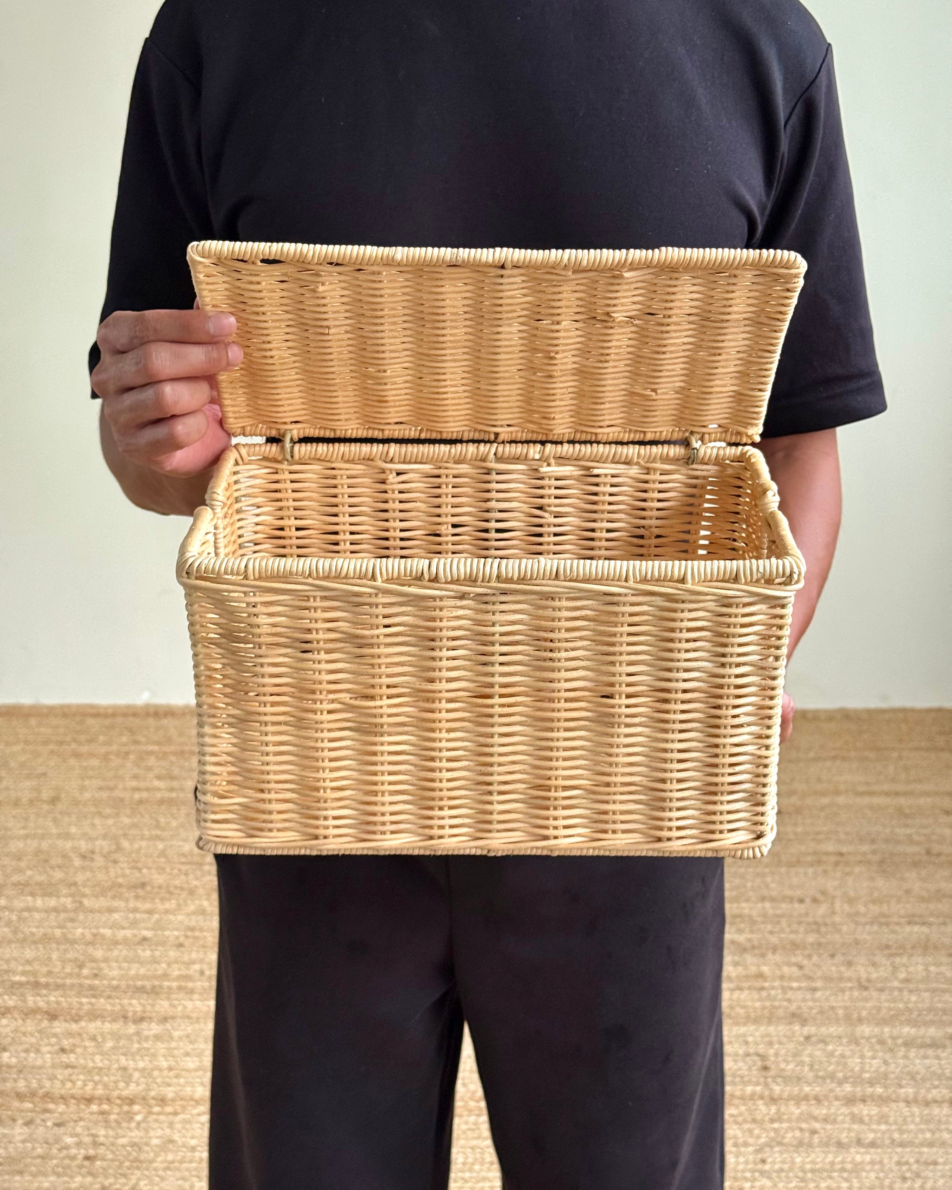 A person wearing a black t-shirt and black pants holds an open natural wicker storage basket against a light beige wall and textured floor.