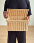 A person wearing a black t-shirt and black pants holds an open natural wicker storage basket against a light beige wall and textured floor.
