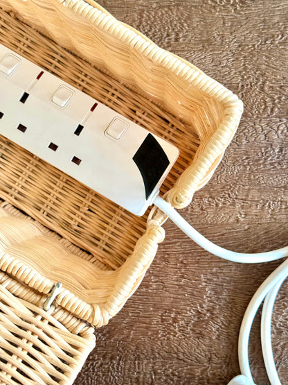A white multi-outlet power strip with a black accent, resting inside a light beige woven wicker basket on a brown textured wooden surface.