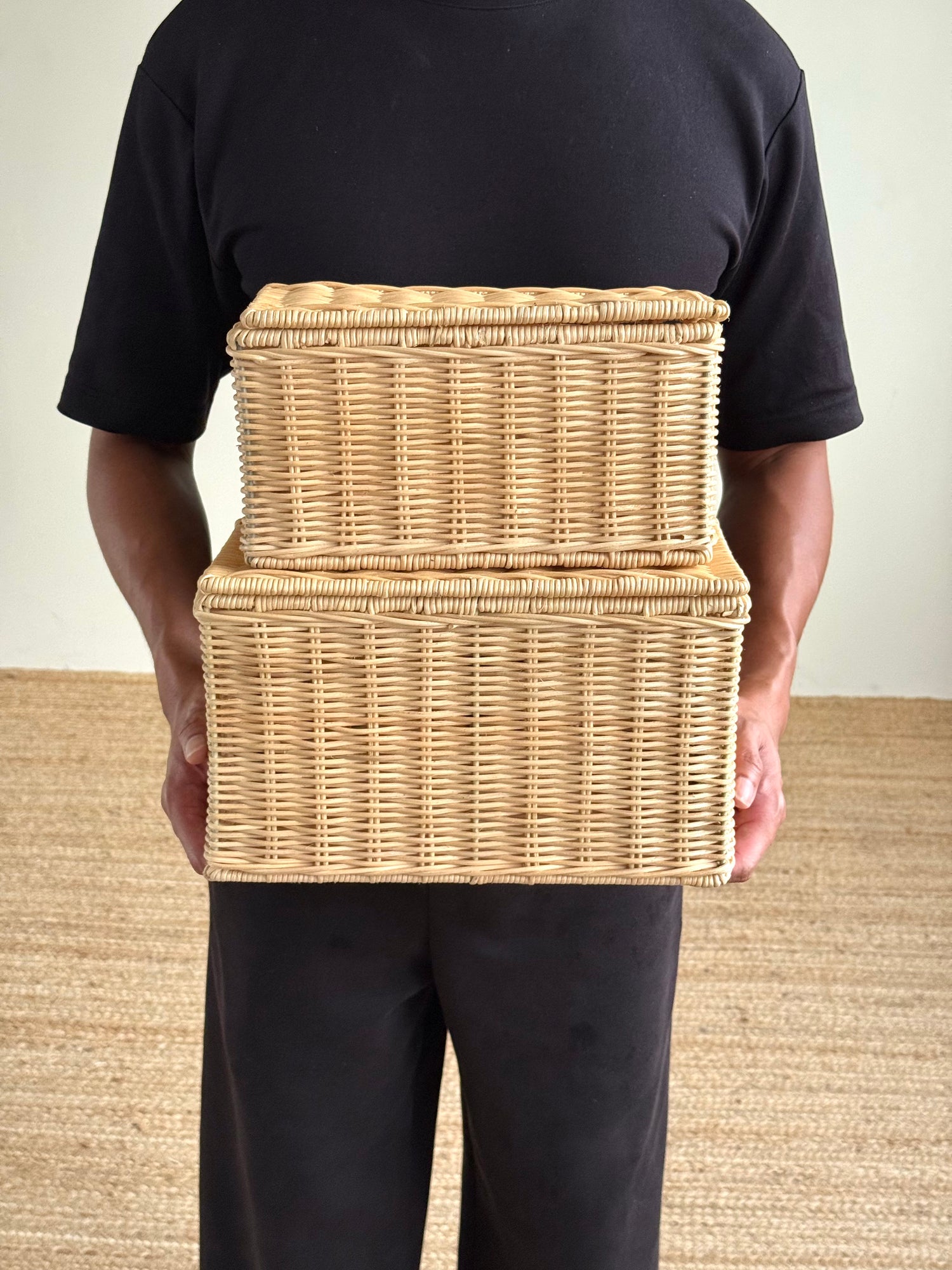 A person wearing a black t-shirt and black pants holds two stacked natural wicker storage baskets against their torso.