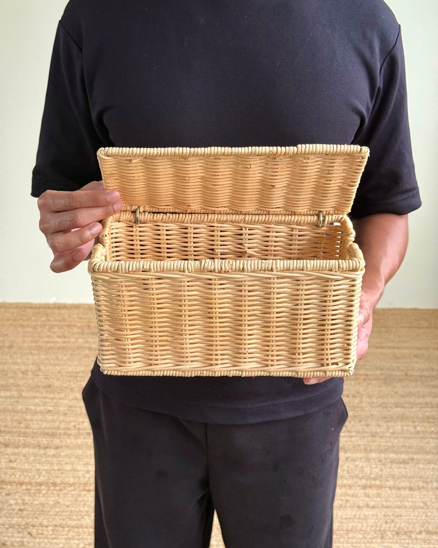 A person wearing a black t-shirt and black pants holds an open natural wicker storage basket against their torso.