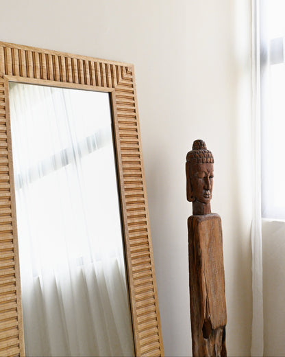 Wooden mirror with wavy frame next to a wooden Buddha statue on a white wall.