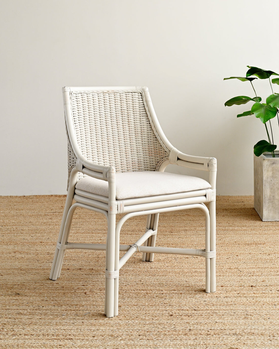 White wicker chair with cushion on a wooden floor next to a potted plant.