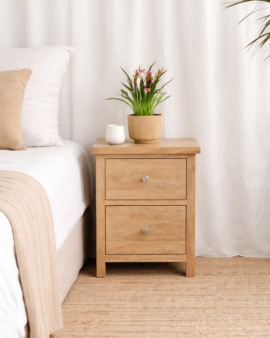 Wooden nightstand with a plant and a cup next to a bed in a bedroom setting.