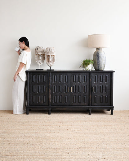 Woman standing next to a black sideboard with decorative items in a minimalistic room.