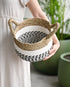 A person holding a round seagrass basket with handles, which is predominantly white with black stripes, displayed against a backdrop of indoor plants.